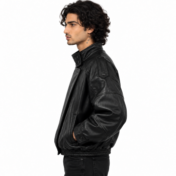 Male model wearing Mizon International men's black oversized genuine leather bomber jacket featuring a high stand-up collar, asymmetrical overlapping front, tonal snap-button details, voluminous sleeves and gathered-looking waistband against a white background.